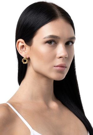 Goldene Creolen mit einem gedrehten Design und strukturierter Oberfläche, präsentiert von einem Modell in einem schlichten weißen Oberteil vor einem weißen Hintergrund.