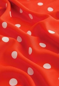 Tessuto rosso con pois bianchi, texture liscia, drappeggiato per mostrare pieghe e profondità. Il motivo presenta pois di diverse dimensioni, equidistanti.