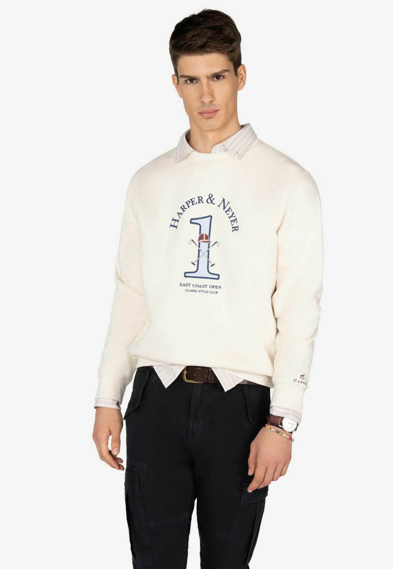 Sudadera de crema clara con un estampado gráfico que presenta "Harper & Never" y el número 1, superpuesta sobre una camisa blanca a rayas y pantalones negros.
