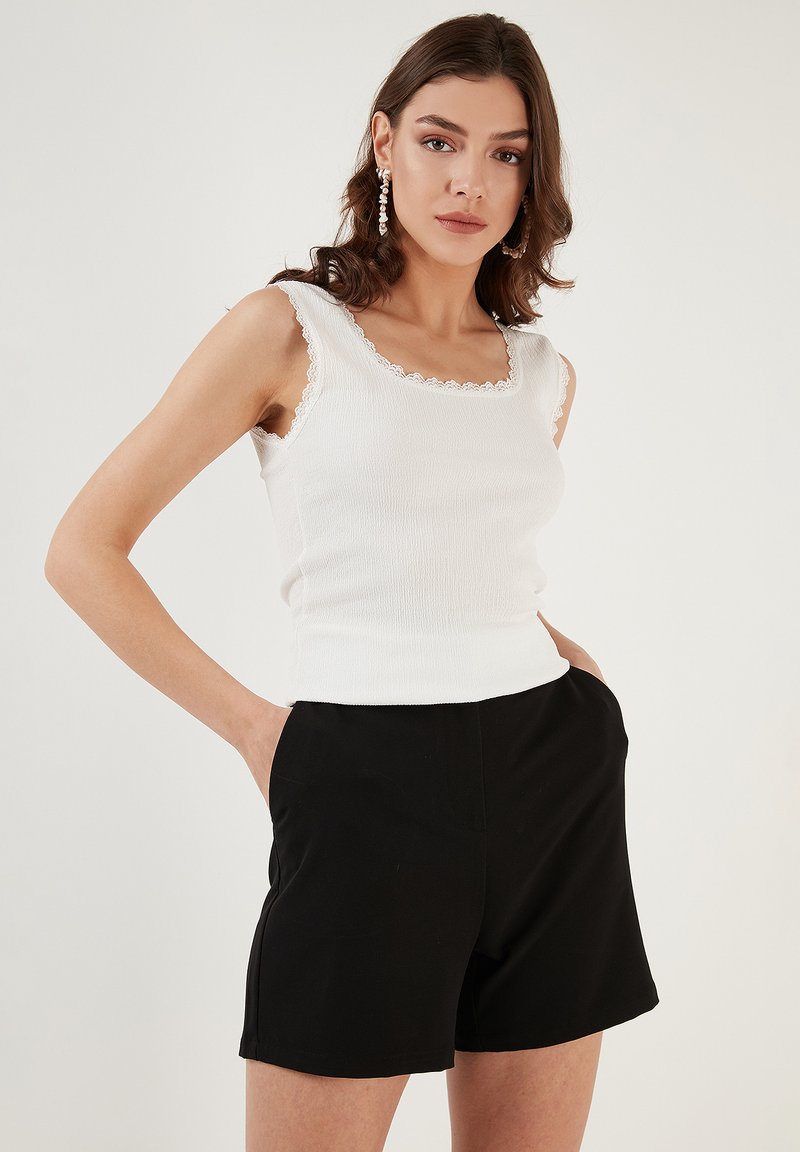 Valge ribaga tank top, mille servas on lainelised pitsid, on koos mustade lühikesed püksidega. Mudel poeb käed taskutesse, näidates riietuse lihtsust.
