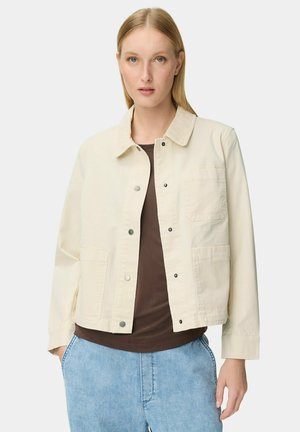 Kobieta o prostych blond włosach, ubrana w beżową zapinaną na guziki kurtkę na ciemnobrązową koszulę i jasnoniebieskie dżinsy, stojąca na tle białego tła.