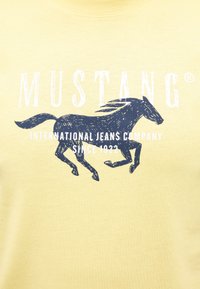 Gelbes Baumwoll-T-Shirt mit einem navyblauen Pferde-Graphic und dem weißen Text "MUSTANG® INTERNATIONAL JEANS COMPANY SINCE 1932".