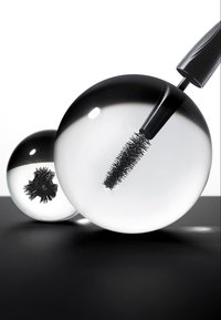 Two transparent spherical lenses magnify a black mascara wand on a black and white surface.