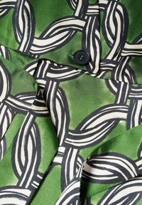 Tissu vert avec un motif de chaîne entrelacée noir et blanc, texture satinée, et un petit bouton noir cousu près d'un pli de col.