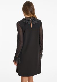 Robe noire avec un design à manches longues transparentes, décolleté froncé et accents en dentelle aux manches. Présente un détail en forme de trou de clé au dos.