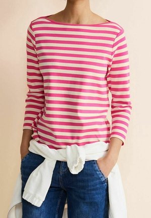 Osoba nosząca różowo-biały pasiasty shirt z długim rękawem, niebieskie dżinsy i biały sweter związany wokół talii, ręce w kieszeniach.