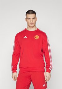 Rode sweatshirt met lange mouwen, witte drie-streep ontwerp langs de schouders, met een Manchester United logo op de borst.
