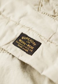 Beżowa tkanina z czarną metką, na której złotymi literami wytłoczono napisy "SUPERDRY" i "WORK-WEAR". Widoczne szwy na materiale.