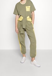 Olivově zelené tričko s žlutou kapsou, doplněné olivově zelenými tepláky se žlutými bočními pruhy. Bílí tenisky dotvářejí celkový vzhled.