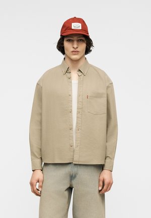 Fiatal személy rozsdavörös sapkában és bézs gombos ingben, fehér pólóval alatta, világos, koptatott farmernadrágban, fehér háttér előtt állva.