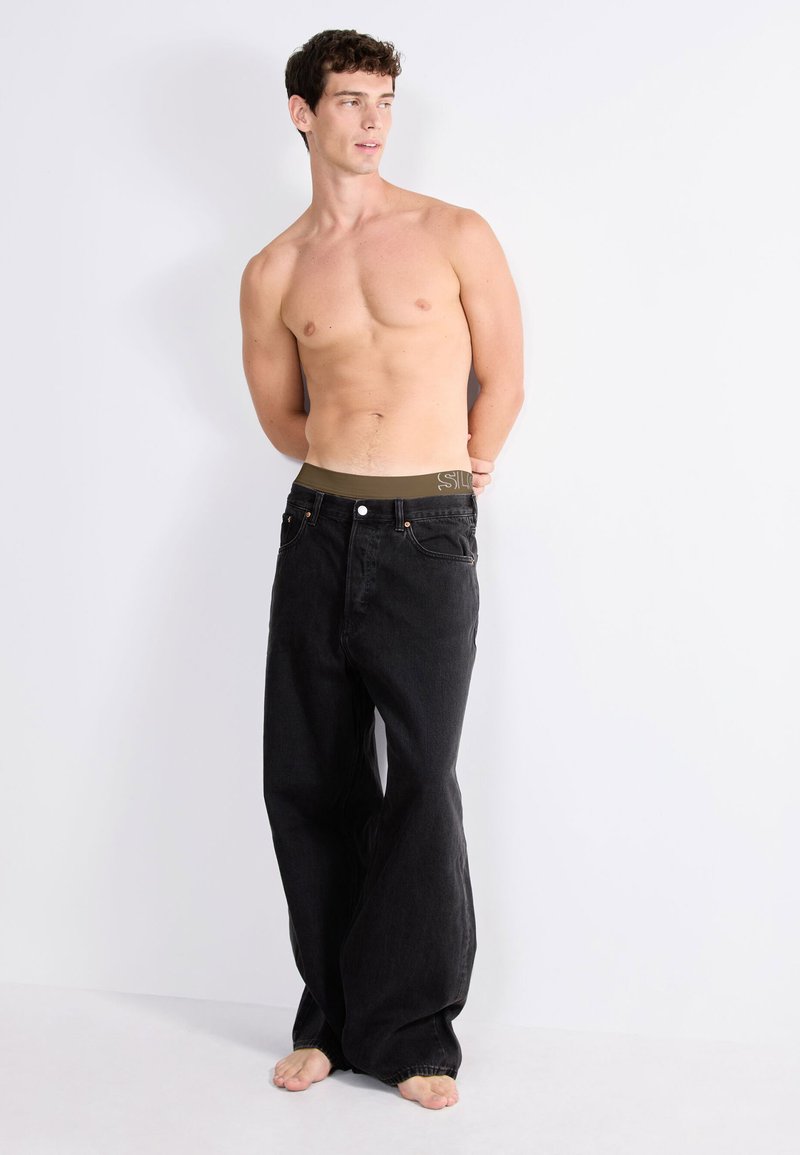 Schwarze Jeans aus Denim mit lockerer Passform und weitem Bein. Der Bund ist olivgrün mit Branding, und der Stoff zeigt eine leichte Textur.