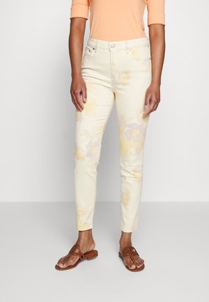 Person trägt hellgelbe, taillierte Hose mit Blumenmuster, pfirsichfarbenes Langarmshirt und braune Sandalen mit offenen Zehen, steht vor einem schlichten Hintergrund.