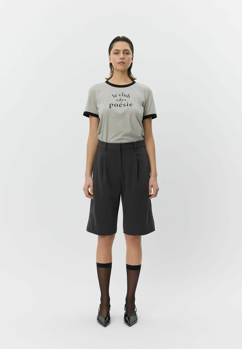 Szara koszulka z czarnym wykończeniem i nadrukiem "le club de poésie", zestawiona z czarnymi, dopasowanymi spodenkami oraz przezroczystymi, czarnymi podkolanówkami, noszona z czółenkami na obcasie.