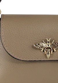Beige leren handtas met een gestructureerde afwerking, voorzien van een gouden bij-embleem op de klepsluiting en contrasterende stiksels langs de randen.
