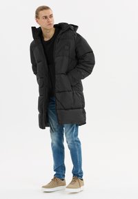 Sort pufferjakke med hætte, med en quiltet design, frontlynlås og sidelommer; båret over en sort top og blå jeans.