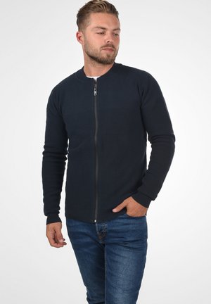 Marineblå sweatshirt med glidelås og rund hals, teksturerte paneler over brystet, ribbestrikkede mansjetter og hem, båret med blå jeans.