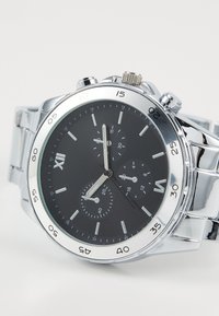 Montre en acier inoxydable argenté avec un cadran noir, dotée d'une lunette chromée, de trois sous-cadrans, d'index horaires argentés et d'un bracelet texturé.