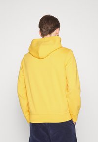 Gul huvtröja med mjuk textur, ribbade muddar och nederkant; har en rundad huva och en enkel, minimalistisk design.