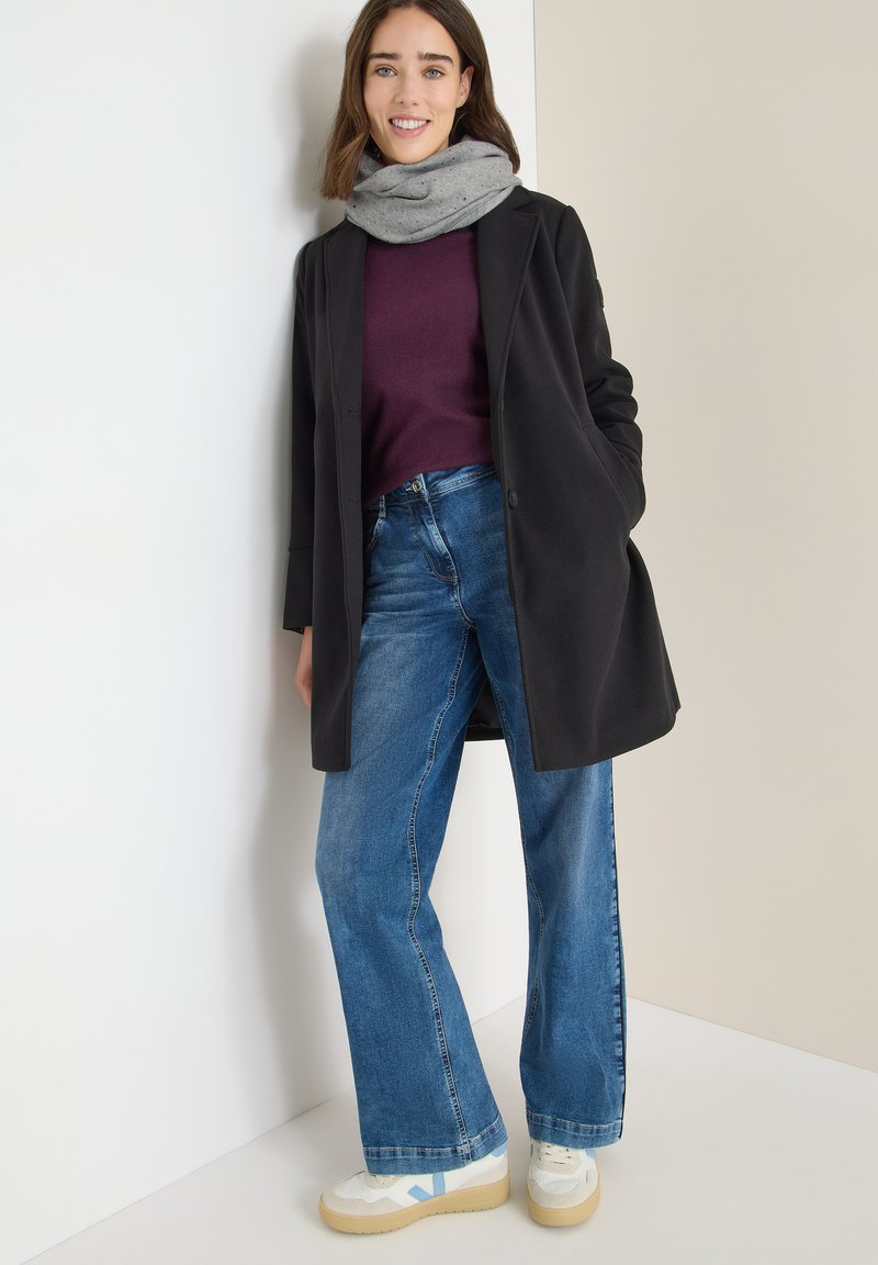 Cappotto nero su un maglione borgogna, abbinato a jeans a gamba larga blu e sneakers bianche con accenti blu. Sciarpa grigia drappeggiata attorno al collo.