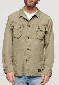 Olijfgroene button-up jas met twee borstzakken, bruine knopen en verstelbare manchetten; gemaakt van zachte, gestructureerde stof.