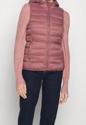 Frau trägt eine mauvefarbene, gesteppte ärmellose Weste mit Reißverschluss über einem rosa langärmeligen Strickpullover und dunkelblauen Jeans.