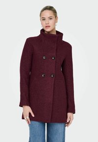 Vīna sarkanā vilnas maisījuma mētelis ar augstu apkakli, dubultā pogu rindas priekšpusi, melnām pogām un piegriezuma siluetu, apvienots ar gaiši zilām džinsu biksēm.