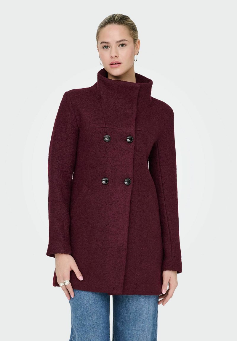 Vīna sarkanā vilnas maisījuma mētelis ar augstu apkakli, dubultā pogu rindas priekšpusi, melnām pogām un piegriezuma siluetu, apvienots ar gaiši zilām džinsu biksēm.