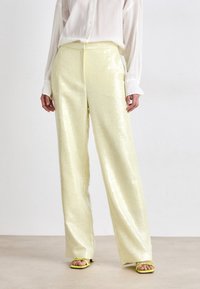Broek in glanzende, lichtgele stof met een gestructureerde afwerking. Losse pasvorm en wijd uitlopende pijpen. Gecombineerd met gele sandalen. Witte blouse zichtbaar.