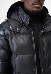 Schwarze Steppjacke aus Leder mit hohem Kragen, Reißverschluss und Knopfdetails. Verfügt über eine glatte Textur und ein gestepptes Design.