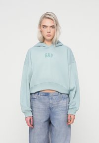 Hoodie cropped di colore azzurro chiaro con logo frontale, caratterizzato da una texture morbida e maniche larghe. Abbinato a jeans in denim di lavaggio chiaro.