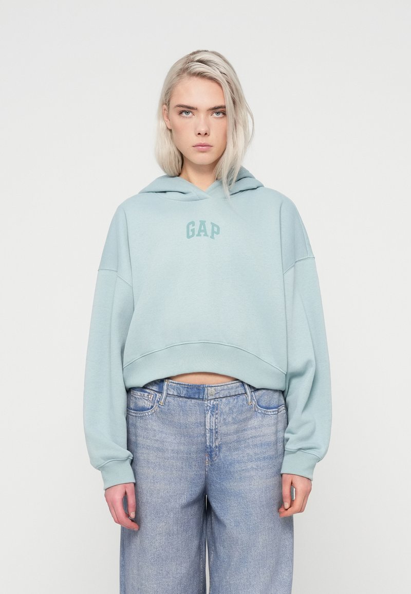 Hoodie cropped di colore azzurro chiaro con logo frontale, caratterizzato da una texture morbida e maniche larghe. Abbinato a jeans in denim di lavaggio chiaro.