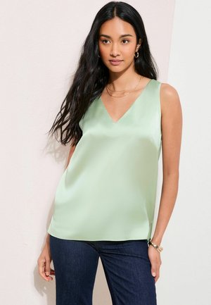 Kobieta o długich ciemnych włosach, ubrana w jasnorozielony, bezrękawnik z dekoltem w serek oraz ciemnoniebieskie dżinsy, stojąca na jasnym tle.