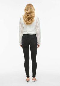 Witte textuurblouse met zigzagpatroon, doorzichtige lange mouwen; gecombineerd met aansluitende zwarte jeans en beige sandalen met bandjes.