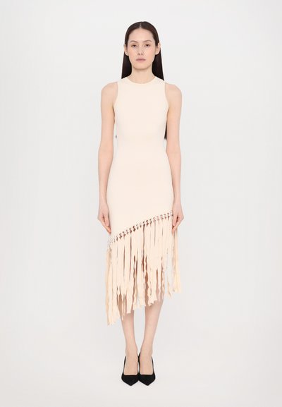 Femme portant une robe midi beige sans manches avec des franges diagonales et des détails en perles, assortie à des escarpins noirs, se tenant sur un fond uni.