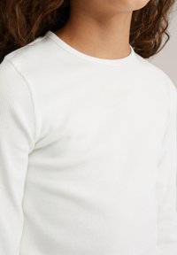 Haut à manches longues blanc à côtes, avec un col rond et une texture douce. Le tissu présente des plis légers et des imperfections subtiles.