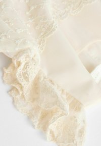 Tissu en dentelle couleur crème avec broderie florale complexe et bords festonnés sur un fond textile doux et transparent.