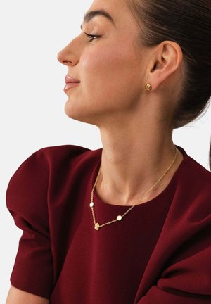 Frau in einem burgunderfarbenen Oberteil, die goldene Steckohrringe und eine filigrane goldene Kettenhalskette mit kleinen geometrischen Anhängern trägt.