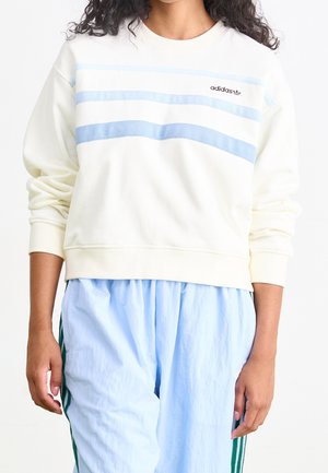 Osoba ubrana w biały sweter Adidas z jasnoniebieskimi paskami oraz niebieskie spodnie z zielonymi paskami po bokach, stojąca na jednolitym tle.