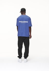Modré bavlněné tričko s bílým nápisem "PEGADOR" na zadní straně, v kombinaci s černými tepláky a bílými teniskami. Jednoduchý design a uvolněný střih.