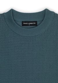 Zbliżenie na turkusowy sweter dzianinowy z prążkowanym okrągłym dekoltem oraz czarną metką "PAKO LORENTE" w rozmiarze M wszytą wewnątrz kołnierza.