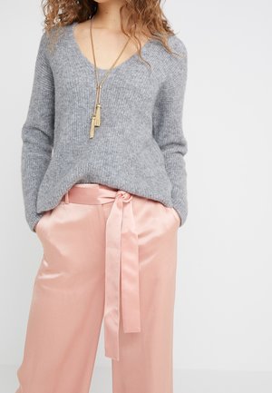 Femme portant un pull en maille gris et un pantalon en satin rose taille haute avec une ceinture nouée, accessoirisé d’un long collier à pampilles dorées.