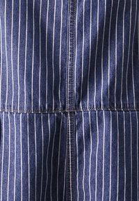 Sininen denimpitoinen kangas, jossa on valkoisia pystysuoria raitoja, näkyvät ompeleet saumoissa ja hieman rypistyvä rakenne vyötäröllä.