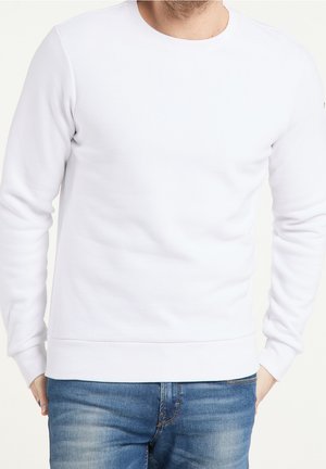 Uomo che indossa una felpa bianca semplice con collo a girocollo e polsini e orlo a coste, abbinata a jeans blu sbiaditi, con le mani parzialmente nelle tasche.