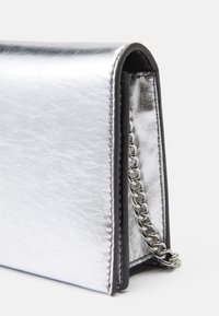 Sudraba metāla clutch soma ar teksturētu virsmu, kas aprīkota ar ķēdes siksnu un minimālistisku dizainu, izceļot slaidas, leņķiskas formas.