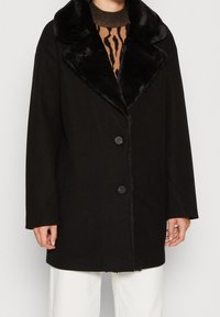 Svart oversized jacka med krage av fuskpäls, med rak snitt, två frambutoner och mjuk textur. Bärs över en mönstrad tröja.