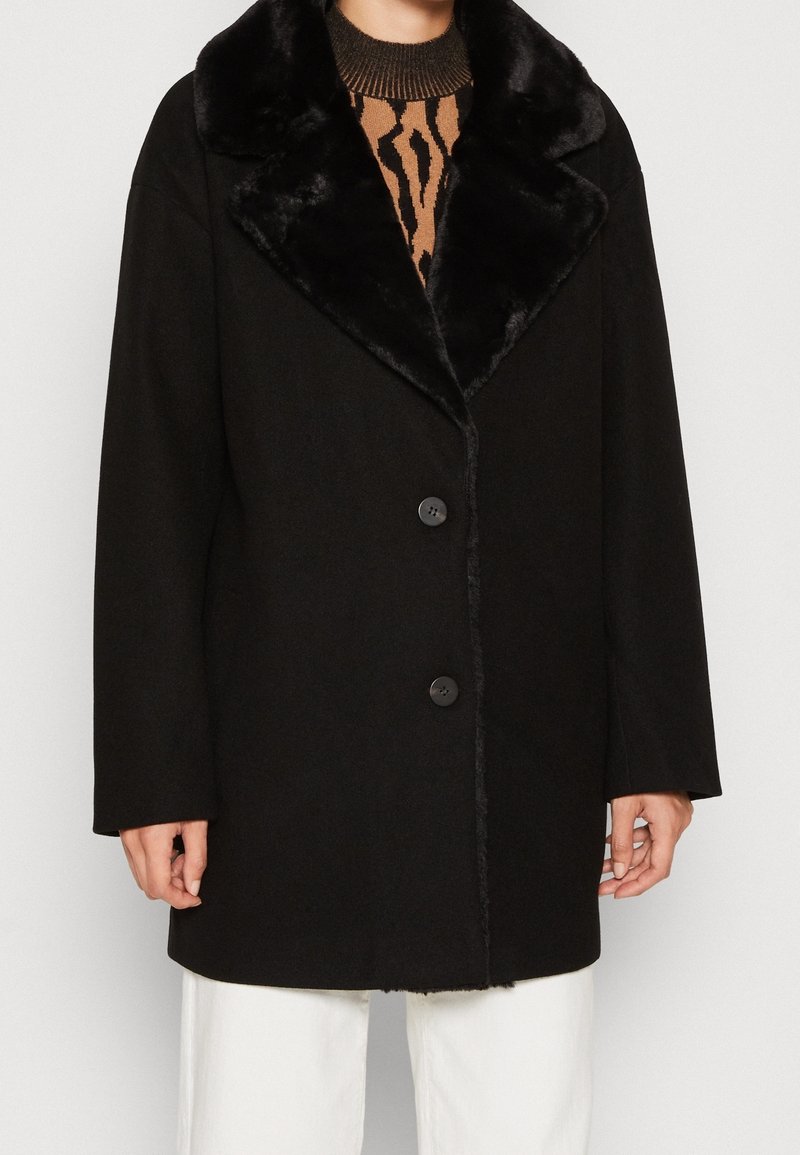 Svart oversized jacka med krage av fuskpäls, med rak snitt, två frambutoner och mjuk textur. Bärs över en mönstrad tröja.
