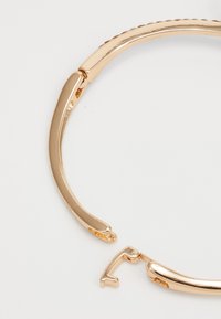 Bracciale in metallo dorato caratterizzato da un design liscio e curvo con un meccanismo di chiusura. La superficie ha una texture lucida con dettagli sottili.