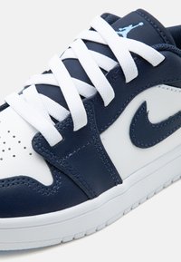 Λευκό και ναυτικό μπλε δερμάτινο sneaker με λευκά κορδόνια, το λογότυπο Nike Swoosh και το λογότυπο Jumpman στη ναυτική γλώσσα, με ορατή λεπτομερή ραφή.