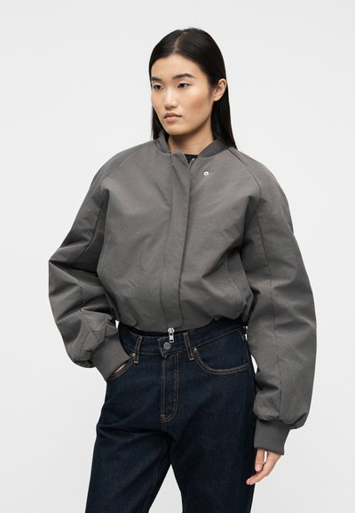 Γυναίκα με ίσια σκουρόχρωμα μαλλιά, που φορά μια γκρι μπουφάν τύπου bomber και σκούρο μπλε τζιν, στέκεται με το ένα χέρι στην τσέπη μπροστά σε απλό φόντο.