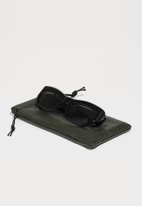 Occhiali da sole neri con montatura quadrata e lenti scure, poggiati su un sacchetto di pelle verde oliva con superficie texturizzata e chiusura a cordoncino.